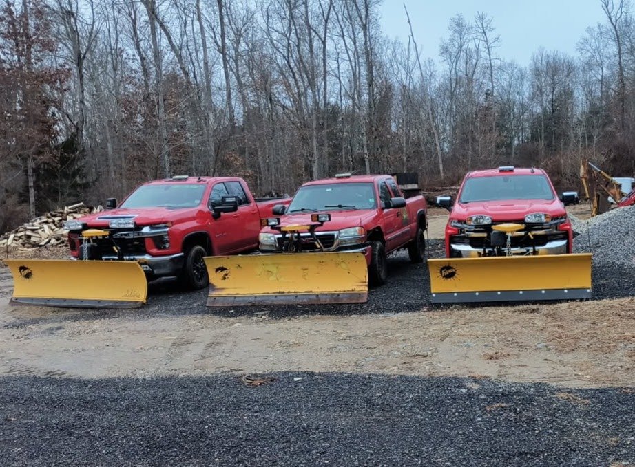 Three red pickup trucks with yellow snow plows, ready for Residential Snow Removal, are parked side by side on a gravel lot in Northeast Connecticut, with trees and stacks of firewood in the background.