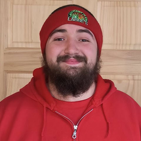 A person is wearing a red beanie and hoodie, standing indoors against a wooden background. Perhaps they just took a break from their free time passion for lawn care with Labbe's Lawn Care Plus.