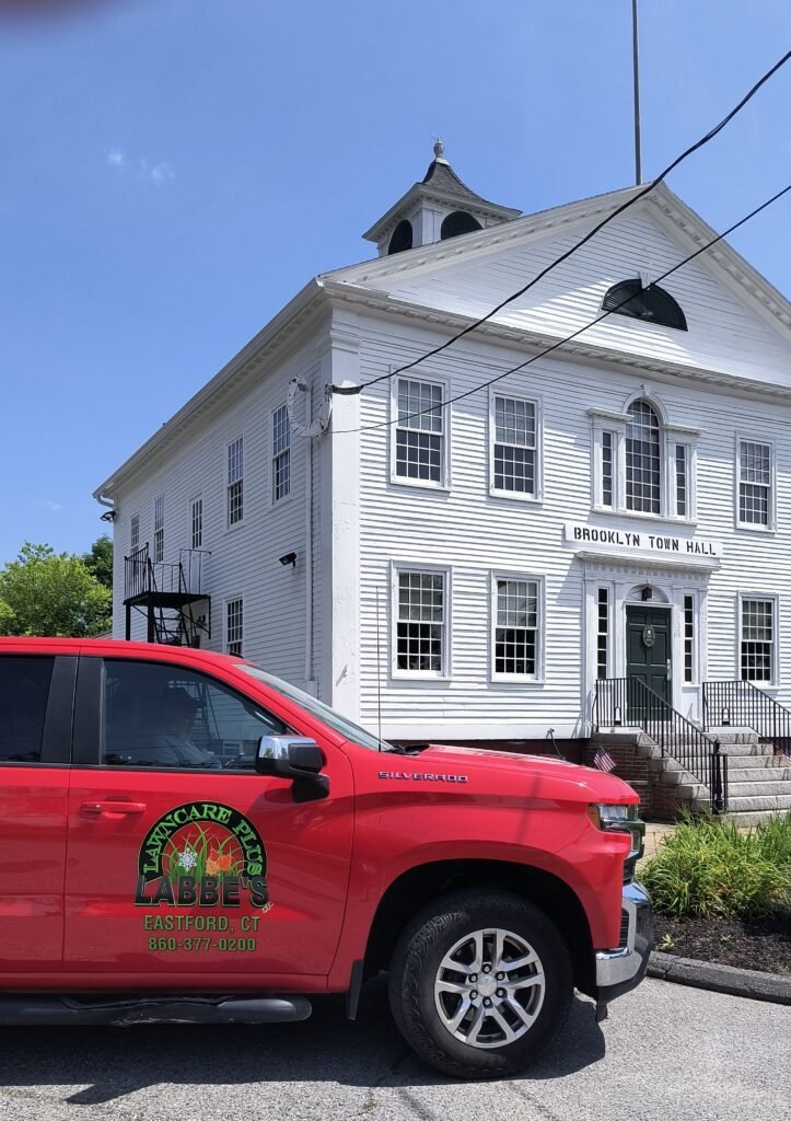 A red landscaping truck from the best company in Brooklyn CT is parked in front of Brooklyn Town Hall, a white, two-story building with black shutters and a cupola.