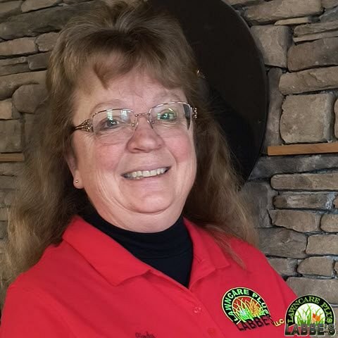 In their free time, a smiling person wearing glasses and a red polo shirt with the Labbe's Lawn Care logo stands proudly in front of a stone wall.