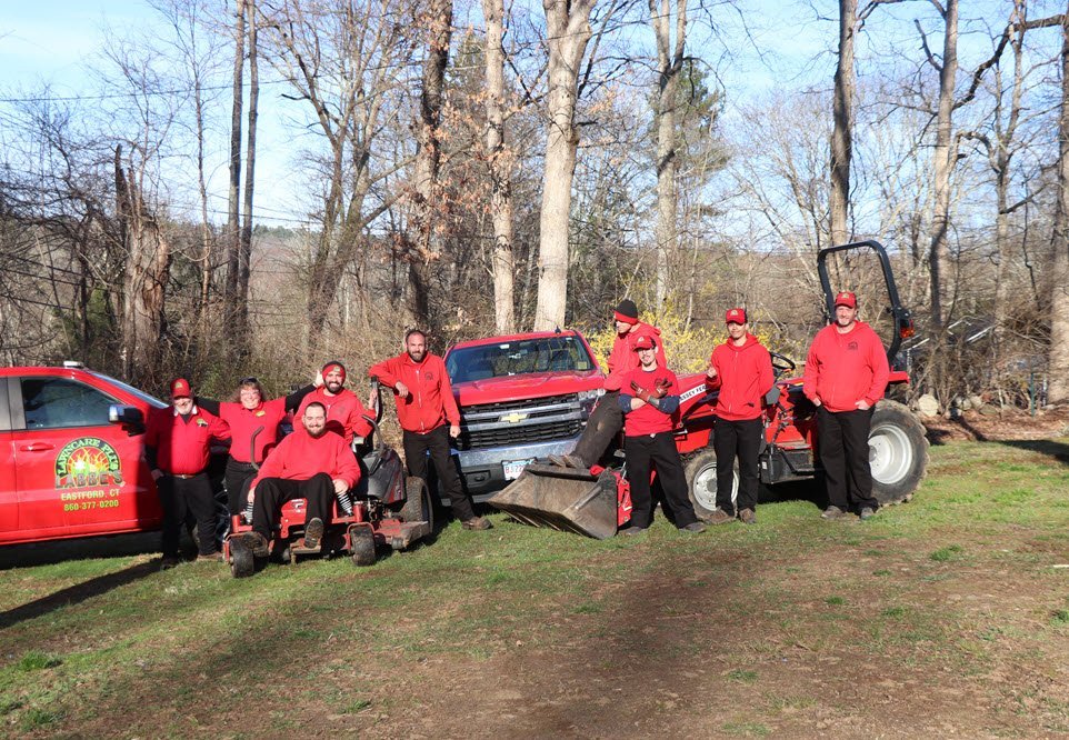 A group of nine people in matching red shirts pose outdoors with two red vehicles and a red tractor on a grassy area surrounded by trees.