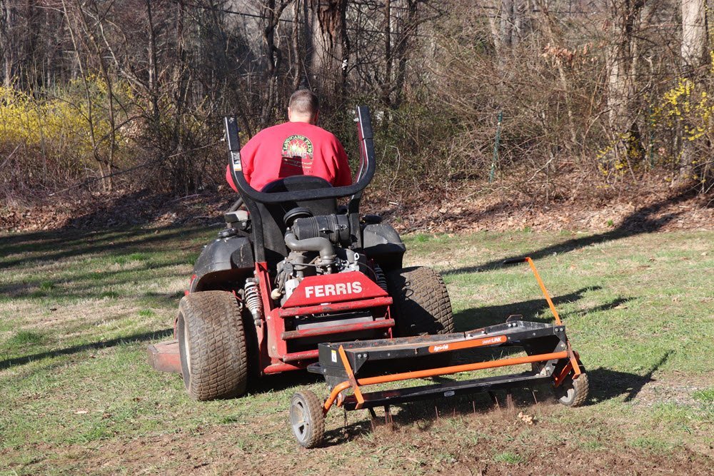 A person performs lawn maintenance by operating a Ferris lawn mower with an attached soil aerator on a grassy area near trees and bushes.