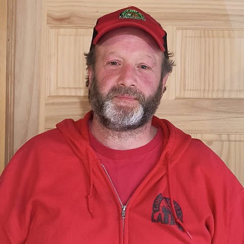 A man with a beard, enjoying his free time, stands in front of a wooden door, wearing a red hat and hoodie.