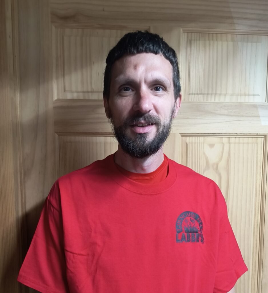 A man with short dark hair and a beard is standing indoors in front of a wooden door, wearing a red T-shirt with a small black logo—ready to spend his free time enjoying his beautiful lawn.