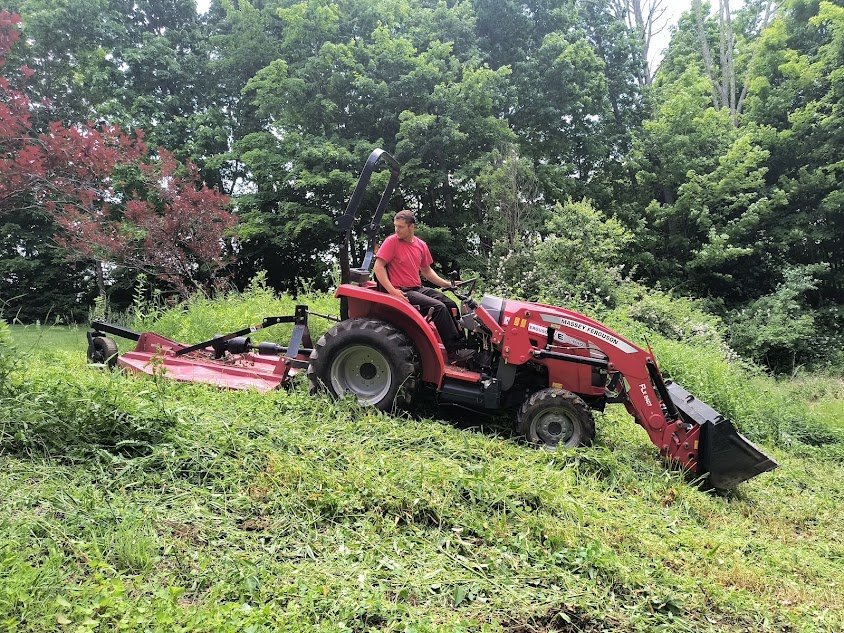 A person operates a red tractor with a mower attachment, reclaiming lawns by trimming overgrown grass on a sloped area surrounded by trees and greenery.