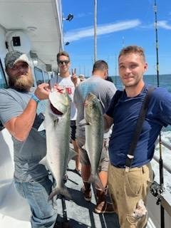 Four men stand on a boat holding large fish they caught; two are displaying their catches while others look on, with the ocean and fishing rods in the background.