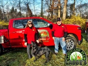 Two people in red shirts stand next to a red truck marked "Abel's" with a black dog in front, in a grassy area with trees in the background.