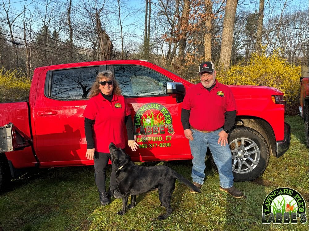 Two people in red shirts stand with a black dog in front of a red truck on a grassy area. Bare trees are in the background.