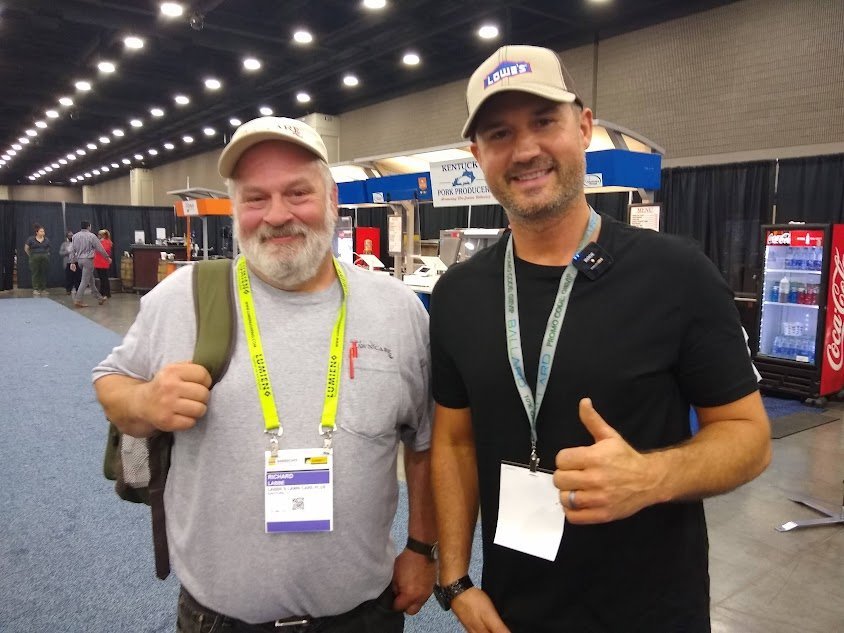 Two men wearing hats and event badges stand indoors at a convention; one smiles with a backpack, the other gives a thumbs up. Exhibition booths are visible in the background.