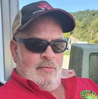 A man with a gray beard wears sunglasses, a red shirt, and a cap while sitting outdoors near a green railing with trees in the background.