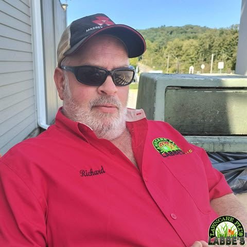 A man with a beard relaxes during his free time, wearing sunglasses, a cap, and a red shirt with "Richard" embroidered on it, as he sits outdoors near a building.