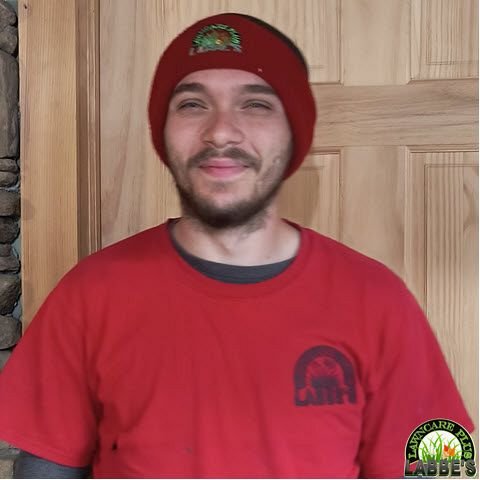 A man wearing a red beanie and red T-shirt stands in front of a wooden door, smiling slightly, enjoying some well-deserved free time after working at Labbe's Lawn Care Plus.