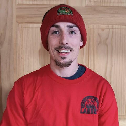 A person smiling, donning a red Labbe's Lawn Care Plus beanie and matching shirt, stands against a wood-paneled backdrop.