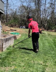 Person in a red shirt and black pants uses a string trimmer to edge grass near a raised garden bed, removing overgrowth and restoring beauty to the sunny backyard.