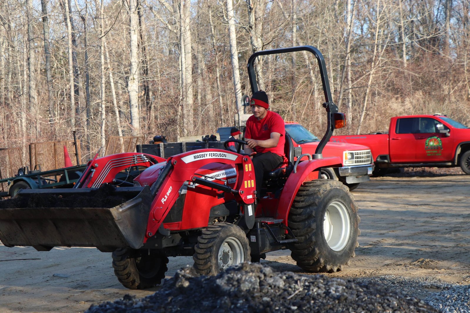 Brooklyn CT A person in a red shirt drives a red Massey Ferguson tractor on a dirt lot, with a red truck and leafless trees in the background.