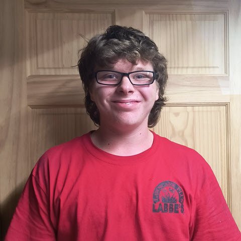 A person with short, curly brown hair and glasses smiles while wearing a red Labbe's t-shirt, standing in front of a wooden door. A person with short, curly brown hair and glasses smiles while wearing a red Labbe's t-shirt, standing in front of a wooden door.