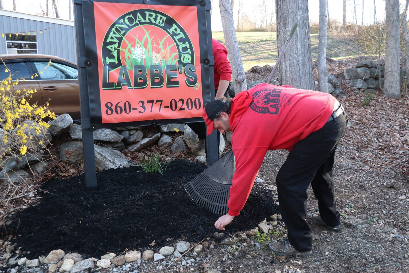 A person in a red sweatshirt spreads mulch with a rake near a "Lawn Care Plus Labbe's" sign and landscaping, showcasing professional mulching services on a sunny day.
