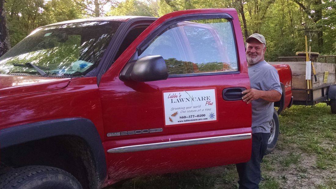 A man stands by the open door of a red pickup truck labeled “Leslie’s Lawn Care, LLC” in a wooded outdoor setting.